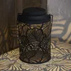 A black metal lantern with a leaf pattern is placed on a patterned surface, casting shadows. Produktbild-Vorschau