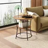 A round side table with two shelves is placed next to a sofa in a living room setting, with a large window showing a winter forest view. Náhľad obrázku produktu