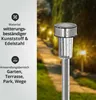 A solar garden stake with water droplets on it, set against a blurred background of a garden path at night with lights. Produktbild-Vorschau