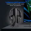 A pair of black over-ear headphones rests on a black charging stand, positioned on a dark surface. A large, out-of-focus television screen with a blue and green abstract image is visible to the right. Produktbild-Vorschau