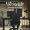 A man is working at a desk with two monitors mounted on adjustable arms. The scene is set in a home office environment with natural light coming from a window. Produktbild-Vorschau