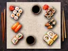 A top-down view of a variety of sushi dishes arranged on a beige placemat with dark brown textured background. Chopsticks and soy sauce dishes are also present. Náhľad obrázku produktu