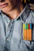 A close-up shot of a person's chest wearing a denim shirt with several colored pens in the pocket. Produktbild-Vorschau