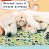 A white puppy sleeps on a green pad with paw print and bone patterns, holding a teddy bear. Produktbild-Vorschau