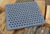 A square cast iron grate with many holes rests on a pile of chopped wood. Voir les images du produit