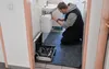 A man is working on plumbing in a bathroom, with a protective mat on the floor. Produktbild-Vorschau