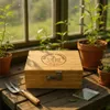 A wooden storage box with a combination lock and engraved logo sits on a wooden surface next to a small gardening tool and potted plants, with a window in the background. Produktbild-Vorschau