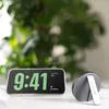 A digital alarm clock displaying the time and date is placed on a white surface, with a charging stand to its right. A blurred green plant is in the background. Anteprima dell’immagine prodotto