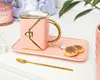 A pink coffee cup and saucer set with a gold handle and trim, accompanied by two cookies and a gold spoon, arranged on a white surface. Produktbild-Vorschau