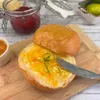 A close-up of a brioche bun split in half, with one half spread with orange marmalade and garnished with lime zest, resting on a wooden cutting board. Jars of jam and limes are visible in the background. Produktbild-Vorschau