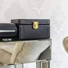 A black leather box with a gold clasp sits on a mirrored surface, with a patterned wallpaper in the background. Produktbild-Vorschau