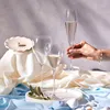 A still life arrangement featuring champagne flutes, a pearl in a shell dish, and draped fabric, with a hand holding one of the flutes. Náhled obrázku produktu