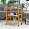 A bamboo serving trolley with three shelves is placed on a carpeted floor next to a gray sofa. The trolley holds bottles of water, books, and headphones. A decorative pillow with a face illustration is on the sofa. Podgląd zdjęcia produktu