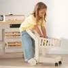 A young girl is playing with a wooden toy shopping cart in front of a toy store counter. Produktbild-Vorschau