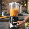 A hand is pressing the power button on a blender filled with an orange smoothie in a kitchen setting. Produktbild-Vorschau