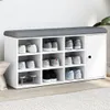A white shoe cabinet with a gray upholstered top, filled with various shoes, placed against a white wall. Náhled obrázku produktu