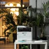 A Brother MFC-L3760CDW multifunction printer is placed on a white table in an office setting, surrounded by lush green plants and a large, ornate chandelier in the background. Produktbild-Vorschau