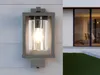 A modern outdoor wall lamp with a clear glass cylinder and a gray metal frame is mounted on a white clapboard wall. To the right, a large window of a modern house reflects the interior. Anteprima dell’immagine prodotto