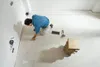 A man is kneeling on the floor and measuring with a tape measure. There are some tools and tiles on the floor. Voir les images du produit