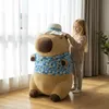 A young girl is adjusting the hat on a large capybara plush toy in a room with sheer curtains and wooden flooring. Produktbild-Vorschau
