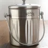 A stainless steel kitchen composter with a handle and lid, placed on a wooden surface. Náhled obrázku produktu