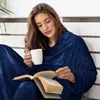 A woman is sitting and reading a book while holding a mug, wrapped in a navy blue blanket with sleeves. Produktbild-Vorschau