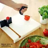 A hand is using a cutting board with a tomato slicer attachment to cut a tomato. Another tomato and a bowl of chopped tomatoes are visible. Produktbild-Vorschau