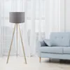 A floor lamp with a gray shade and a wooden tripod base stands next to a light blue sofa in a room with sheer white curtains. Anteprima dell’immagine prodotto