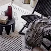 A black woven chair with a black and white patterned throw blanket draped over it, placed on a rug with a black and white geometric pattern. A small dark side table with a decorative box and a dark red vase with dried flowers is next to the chair. Produktbild-Vorschau