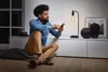 A man is sitting on the floor in a living room, using his smartphone. A table lamp is on in the background. Produktbild-Vorschau