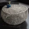 A round, silver coffee table with an intricate, organic design sits on a dark floor, with a dark gray sofa visible in the background. Two small blue vases with delicate white flowers are placed on the table.