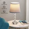 A table lamp with a fabric shade and crystal accents sits on a marble-top side table next to a teal armchair. A smartphone is charging via the lamp's USB port. Produktbild-Vorschau