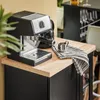 An espresso machine and a small metal pitcher are placed on a wooden countertop, next to a black cabinet. Produktbild-Vorschau
