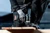 A person wearing work clothes and gloves is using a cordless drill to drill into a piece of wood. Anteprima dell’immagine prodotto