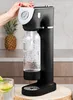 A woman's hand is shown operating a black soda maker with a clear bottle filled with bubbly water. A pineapple is visible in the background, and a glass with lime slices is in the foreground. Produktbild-Vorschau