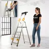 A woman stands next to a KADAX aluminum ladder in a room with white walls and a large window. Podgląd zdjęcia produktu