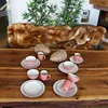 A table setting with pink and white dishes, decorative pinecones, and a fur throw on a sofa in the background. Produktbild-Vorschau