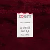 A close-up of a fabric label with care instructions and brand information, attached to a dark red fuzzy fabric. Produktbild-Vorschau