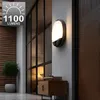 An outdoor wall lamp with a motion sensor is mounted on a white wall next to a wooden door. The lamp is illuminated, casting a warm glow. In the background, a large window with dark frames reveals an interior with a lamp and a wooden cabinet with books. Voir les images du produit