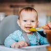 A baby is being fed by a spoon while sitting in a high chair. Anteprima dell’immagine prodotto