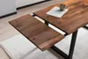 A close-up view of a wooden dining table with an extension leaf pulled out, showing its natural wood grain and a dark metal frame. Produktbild-Vorschau