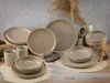 A collection of ceramic tableware, including plates, bowls, and mugs, arranged on a wooden surface with dried grass accents. Produktbild-Vorschau