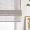 A close-up view of a gray Roman blind with a subtle dotted pattern, hanging in front of a window. A framed picture with colorful flowers and a green plant are visible to the right. Produktbild-Vorschau