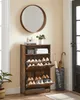 A narrow, dark wood shoe cabinet with two flip-down drawers is shown in a hallway. A round mirror hangs on the wall above the cabinet. An umbrella stand with two umbrellas sits to the right of the cabinet. Náhled obrázku produktu