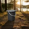 A dark gray camping toilet bucket with a lid and handle sits on the ground in a forest clearing, with a tent and lake visible in the background during sunset. Anteprima dell’immagine prodotto