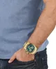 A person's arm and torso are visible, wearing a blue t-shirt and jeans. A gold watch with a green face is on the person's wrist. Náhled obrázku produktu