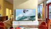 A modern living room with a large TV displaying a surfing scene, two orange accent chairs, and large windows. Produktbild-Vorschau