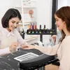 A nail technician is performing a manicure on a client in a salon setting. The image shows a professional salon environment with various nail care products and equipment visible. Voir les images du produit