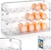 A clear acrylic egg holder filled with eggs, shown from a slightly elevated front-left angle. A smaller inset image shows a label with branding and quantity. Náhled obrázku produktu