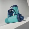 Three turquoise water bottles with dark blue lids are stacked on a light gray surface with a shadow cast by a diagonal line. Podgląd zdjęcia produktu